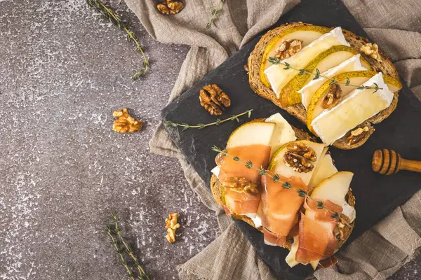 Armut, peynir, fındık, bal, kavrulmuş dilimler ve tahıllı ekmeğin üzerinde bir dal biberiye, sağdaki taş tepside, koyu beton bir masada, solda küçük bir fotokopi alanı olan, düz bir yakın plan. Sağlıklı yeme kavramı.