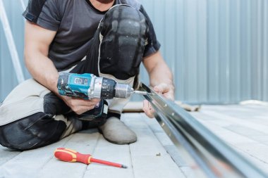 Yazın arka bahçedeki metal barakaya yerleştirmek için çatı pervazını tornavida ve cıvatalarla birbirine bağlayan tanınmayacak kadar olgun beyaz bir adam. Alt taraf görünümü kapalı. Metal dökme konsepti, takım çalışması zamanı, teknoloji kullanımı.