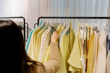 Arkadan duran ve bahar günü bir butikte sarı tereyağı renginde bir yaz tişörtü seçen güzel beyaz bir esmer kızın portresi. Alışveriş zamanını hesapla, kıyafet seç..