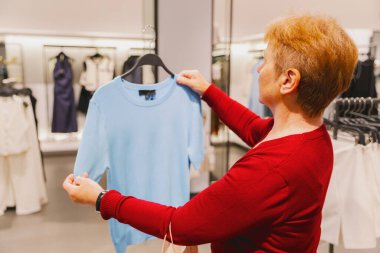 Kırmızı kazaklı, güzel, beyaz, yaşlı bir kadının göğsüne portre, mavi tişörtlü, bir bahar günü bir mağazada duran, seçici bir odak ile yan görüntü. Konsept zaman alışverişi, emeklilik yaşam tarzı, giysi seçimi