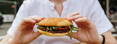 Beyaz tişörtlü, tanınmayan beyaz tenli genç bir adam, iki eliyle meze ve salatalı bir burger tutuyor, bir yaz günü terasta bir masada oturuyor, çok yakından bakıyor. Zaman, fast food demektir..