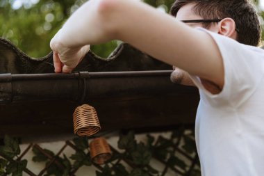 Beyaz tişörtlü, beyaz tenli, esmer bir gencin omuz hizasında portresi. Arka bahçedeki bahçe evinin çatısına çelenk asıyor. Bir bahar günü.