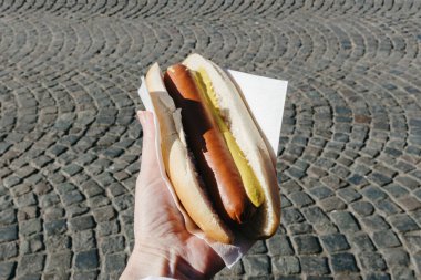 Güneşli bir bahar gününde Brüksel 'in arka planına karşı yürüyen kahverengi ceketli genç bir beyaz adamın elinde mayonez damlalarıyla birlikte güzel kızarmış sosis manzarası. Fast food kavramı