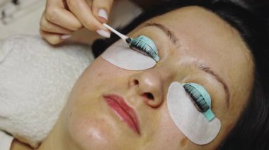 Sağ gözünün kirpiklerini silikon bir formda kaplayan, pamuklu çubukla kahverengi boyanın kalıntılarını temizleyen, kozmetoloji masasında uzanan bir müşterinin elleri.