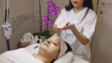 Genç bir kozmetik uzmanı kız, ellerini kullanarak yüzüne masaj maskesini sürüyor. Güzellik salonunda, yakın çekimde gözleri kapalı bir şekilde uzanan bir müşterinin hareketlerini okşuyor. Terapötik yüz temizleme
