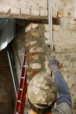Genç beyaz bir adam taze çimentoya tuğla döker, onu spatulayla kaplar ve gün boyunca eski bir evin girişinde dikilir..
