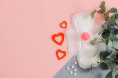 Bir adet pembe virüs topu, üç tahta kalp, beyaz haplar ve okaliptüs dalları sağda gri-pembe bir arka planda uzanıyor..