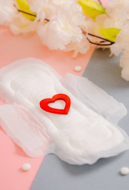 Bir açık kadın yastığı, kırmızı ahşap bir kalple, beyaz tabletlerle ve çiçek açan elma dallarıyla gri-pembe arka planda, seçici bir odak noktasıyla yakın çekimde uzanır..