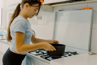 Güzel bir beyaz kızın portresi, Cadılar Bayramı pastasını pişirmek için gaz sobasının üzerine siyah bir tencere koyuyor sonbahar günü mutfakta dikilirken, yakın çekimde. Yemek yapmak için zaman kavramı, tatil hazırlığı.