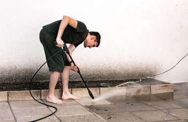 Bir bahar günü arka bahçesinde dikilirken basınçlı çamaşır makinesiyle kirli bahçe fayanslarını yıkayan beyaz bir adamın tam boy portresi. Teknoloji kullanarak bahar çalışma zamanı kavramı.