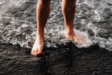 Genç bir beyaz adam dizlerinin altında bir kaldırım taşı üzerinde duruyor, Atlantik Okyanusu 'nun köpüren bir dalgası olan siyah volkanik plajda, sonbahar günbatımı akşamında, yakın çekimde. Konsept: tatil izni, tedavi amaçlı rahatlama.