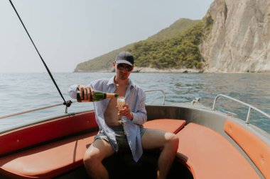 Şapkalı genç beyaz bir adamın portresi, düğmeleri iliklenmemiş bir gömlekle turuncu bir teknenin arkasında oturuyor ve güneşli bir yaz gününde denizde yelken açarken mutlu bir gülümsemeyle bardağa şampanya dolduruyor..