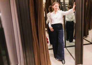 Güzel, beyaz, olgun bir kadının tam boy portresi soyunma odasına bakıyor ve bahar günü alışveriş merkezinde yeni kıyafetlerini gösteriyor. Yan manzara, yakın çekim. Konsept, alışveriş zamanı, mutlu kadın.