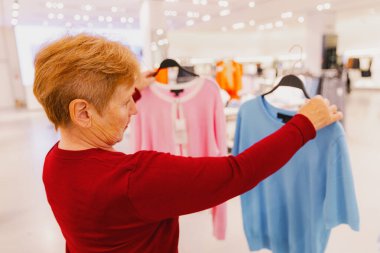 Beyaz, kırmızı kazaklı güzel bir kadının göğüs hizasında portresi. Mavi tişört ve pembe hırka arasında seçim yapıyor. Bahar günü dükkânda duruyor. Konsept zaman alışverişi, emeklilik yaşam tarzı, seçim