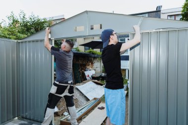 Yazın metal bir barakada çatı kısımlarını birleştiren iki beyaz genç adam, seçici bir odak ile yan görüş. Teknoloji ve zaman takım çalışmasını kullanarak, konsept metal dökümü montajı.