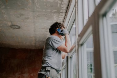 Gri üniformalı, kıvırcık saçlı, genç bir beyaz adam, tadilatın devam ettiği bir evin iskelesinde yan yana duruyor ve yakın çekim sırasında cep telefonuyla konuşuyor. Ev yenileme konsepti, pencere