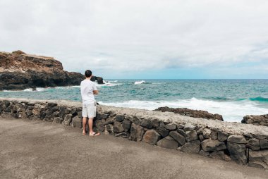 Bulutlu bir sonbahar gününde Atlantik Okyanusu 'ndaki bir adanın kuzey kıyısındaki kayalara çarpan dalgaları izleyen beyaz bir adamın tam boy portresi. Geniş açılı yan görüş. Kavram: rahatlatıcı