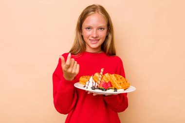 Kafkasyalı küçük bir kız bej arka planda izole edilmiş bir waffle tutuyor. Sanki seni davet ediyormuş gibi parmağıyla işaret ediyor..