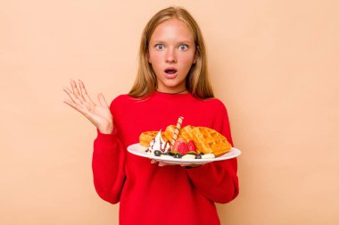 Kafkasyalı küçük bir kız bej arka planda izole edilmiş waffle tutuyor. Şaşırmış ve şok olmuş..