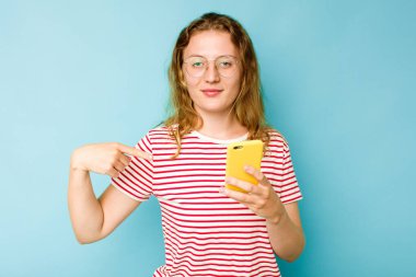 Elinde cep telefonu olan beyaz bir kadın mavi arka planda tek başına gömlek fotokopi alanını işaret ediyor, gururlu ve kendinden emin.