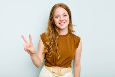 Mavi arka planda izole edilmiş genç beyaz kadın zafer işareti gösteriyor ve gülümsüyor..