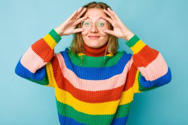 Mavi arka planda izole edilmiş genç beyaz kadın bir başarı fırsatı bulmak için gözlerini açık tutuyor..