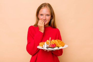 Elinde bej arka planda izole edilmiş waffle tutan küçük beyaz kız tırnaklarını yiyor, gergin ve çok endişeli..