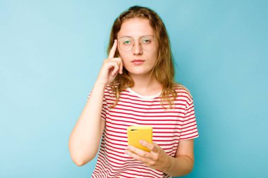 Elinde cep telefonu olan beyaz bir kadın mavi arka planda izole edilmiş. Şakağı işaret ediyor, düşünüyor ve bir göreve odaklanıyor..