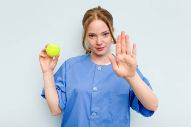 Beyaz tenli genç fizyoterapist elinde mavi arka planda izole edilmiş bir tenis topu tutuyor..