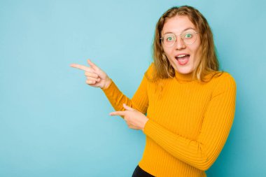 Mavi arka planda izole edilmiş genç beyaz kadın işaret parmağıyla işaret ederek heyecanlanıyor..