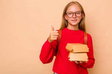 Bej arka planda izole edilmiş hamburgerleri tutan beyaz kız gülümsüyor ve başparmağını kaldırıyor.