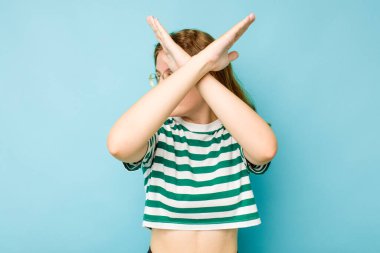 Mavi arka planda izole edilmiş genç beyaz kadın iki kolu çapraz tutuyor, inkar kavramı.