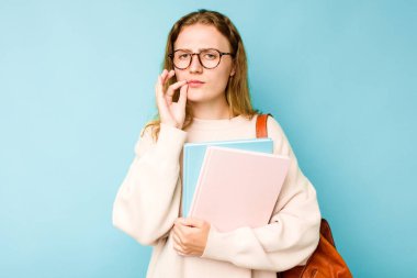 Genç beyaz bir kadın öğrenci mavi arka planda izole edilmiş, parmakları dudaklarında bir sır saklıyor..