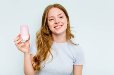 Mavi arka planda deodorant tutan genç beyaz kadın mutlu, gülümseyen ve neşeli..