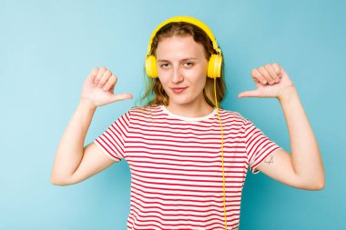 Mavi arka planda izole edilmiş kulaklık takan genç beyaz kadın gururlu ve kendinden emin hissediyor, örnek olarak.