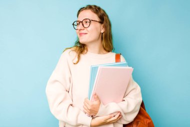 Mavi arka planda izole edilmiş genç beyaz kadın hedeflere ve amaçlara ulaşma hayali kuruyor.