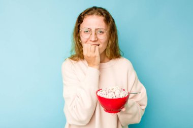 Genç, beyaz bir kadın elinde bir kase mısır gevreği tutuyor. Mavi arka planda izole edilmiş, tırnaklarını yiyor. Gergin ve endişeli..