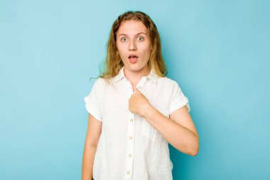 Mavi arka planda izole edilmiş genç beyaz kadın parmağını göstererek şaşırdı, geniş bir gülümsemeyle..