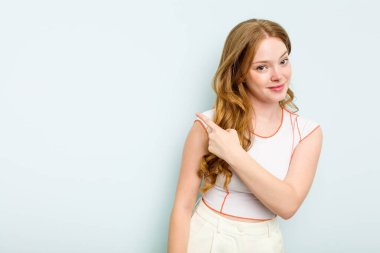 Mavi arka planda izole edilmiş genç beyaz kadın gülümsüyor ve boş alanda bir şeyler gösteriyor..