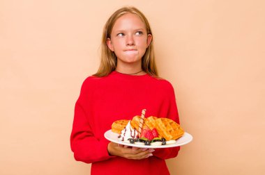 Kafkasyalı küçük bir kız bej arka planda izole edilmiş waffle tutuyor. Kafası karışmış, kuşkulu ve güvensiz hissediyor..
