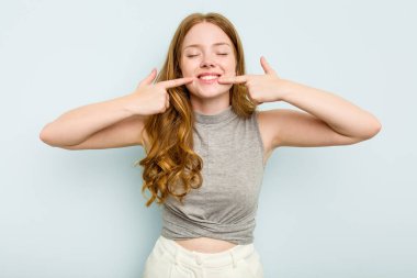 Mavi arka planda izole edilmiş genç beyaz kadın gülümsüyor, parmaklarını ağıza doğrultuyor..