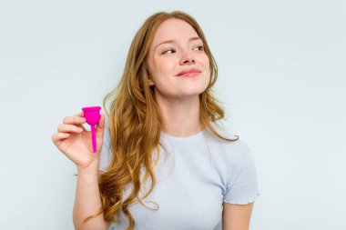 Adet bardağını elinde tutan beyaz bir kadın mavi arka planda izole bir şekilde amaçlarına ve amaçlarına ulaşma hayali kuruyor.