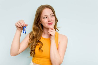 Young caucasian woman holding home keys isolated on blue background looking sideways with doubtful and skeptical expression.