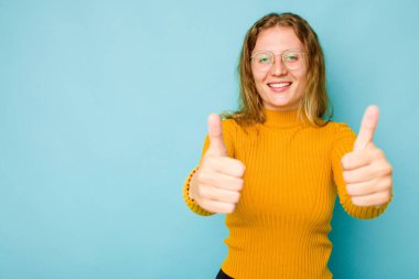 Mavi arka planda izole edilmiş genç beyaz kadın gülümsüyor ve başparmağını kaldırıyor.