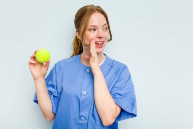 Beyaz tenli genç fizyoterapist elinde mavi arka planda izole edilmiş bir tenis topu tutarken gizli bir sıcak fren haberinin olduğunu söylüyor.