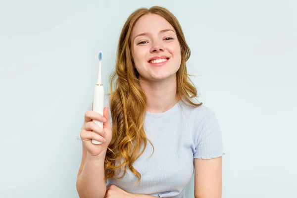 青い背景で孤立した電動歯ブラシを持っている若い白人女性笑いと楽しみを持っている — ストック写真