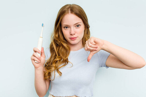 Young caucasian woman holding electric toothbrush isolated on blue background showing a dislike gesture, thumbs down. Disagreement concept.