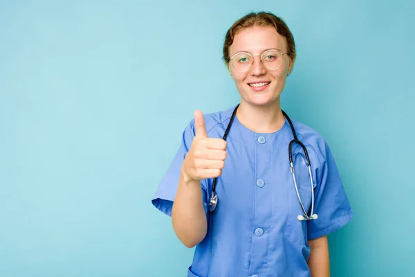 若いです看護師Caucasian女性隔離上の青の背景笑顔と上げ親指アップ — ストック写真