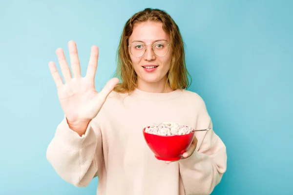 年轻的高加索女人拿着一碗谷物 蓝色背景隔离 笑容满面 带着五号手指 — 图库照片
