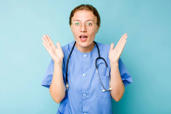 年轻的护士高加索女人被蓝色背景隔离 感到惊讶和震惊 — 图库照片